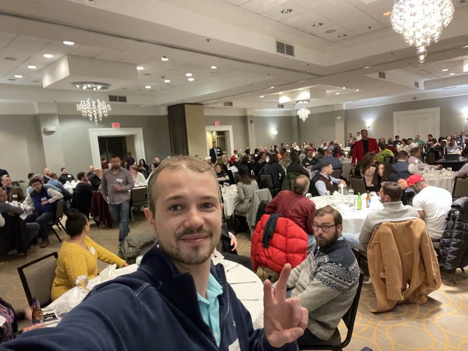 Large networking event or real estate meeting in a banquet hall, with many people seated at round tables. A man in the foreground takes a selfie while flashing a peace sign. Chandeliers and formal décor are visible in the background.